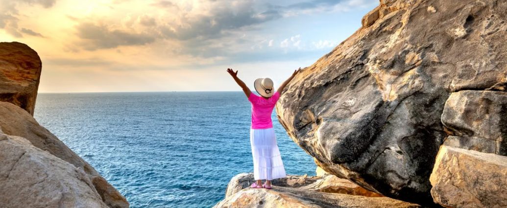 A woman stands on a rocky cliff with arms raised, enjoying the scenic ocean and sunset view.