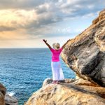 A woman stands on a rocky cliff with arms raised, enjoying the scenic ocean and sunset view.